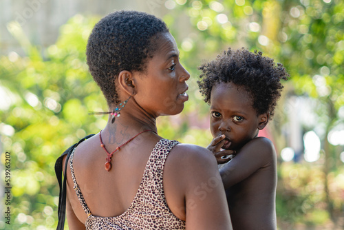 Imagen horizontal de una mujer africana de espalda cargando a su pequeño hijo al aire libre en un día de verano. 