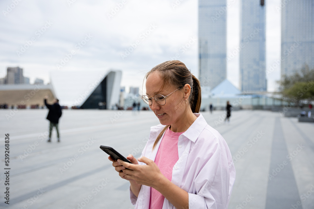 an Asian woman with glasses looks at her phone and smiles in the business district of the city