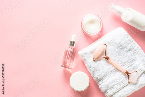 Natural cosmetics on pink. Skin care product, cream, soap serum, jade roller and white towel. Flat lay image with copy space.