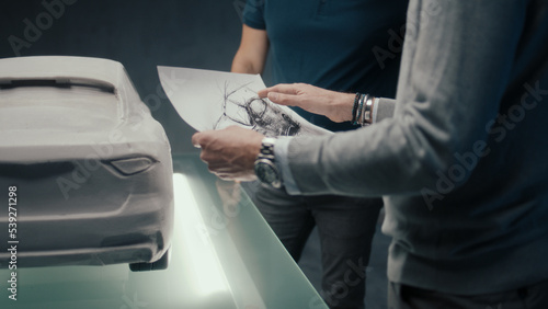 Experienced car designers and developers discuss the design of the car while standing infront of the sculpture of prototype car. Using the pencil sketches in a high tech car manufacturing company.