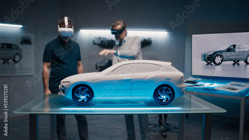 Automotive professional engineers with VR headset and joysticks talk about vehicle production while standing infront of a prototypecar model in a high tech office. Car design analysis and improvement.