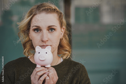 blond young negative thinking woman with depressed look holding pink piggy bank infront of her head while being scary & worried about future and her savings