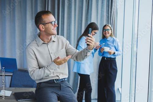 Smiling young male manager in glasses talking on the phone while standing in the office with colleagues in the background. A working day in a modern office with large windows