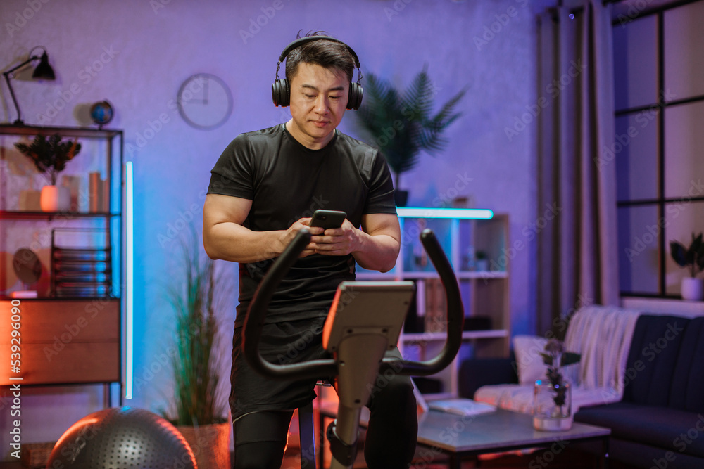 Healthy fit smiling asian man training at home on exercise static bike during workout holding phone, listening music with headphones for motivation. Male healthy weekly habits app. Healthy fit smiling asian man training at home on exercise static bike during workout holding phone, listening music with headphones for motivation. Male healthy weekly habits app.