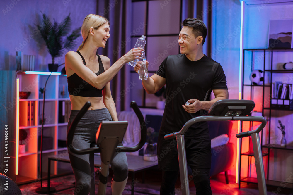 Blond caucasian sports women in sportswear cycling bike and fit asian man running on treadmill while at home at night clinking water bottles, cheers celebrating good results. Blond caucasian sports women in sportswear cycling bike and fit asian man running on treadmill while at home at night clinking water bottles, cheers celebrating good results.