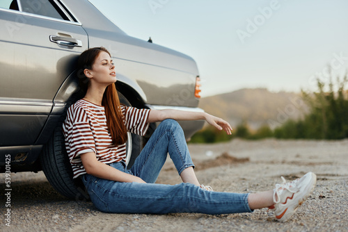 A young woman sits on the ground near her car on the side of the road and looks at the sunset © SHOTPRIME STUDIO