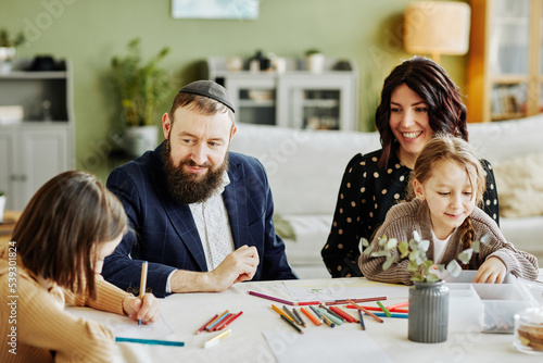 Portrait of modern jewish family drawing together while sitting at table , focus on smiling father wearing kippah