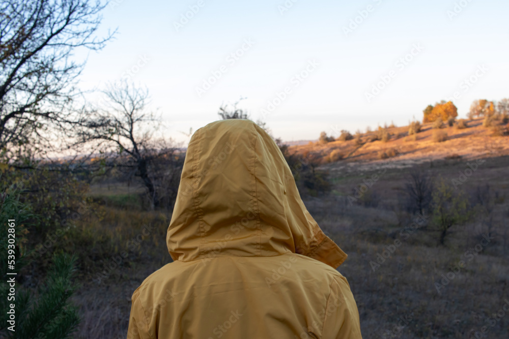 A woman in a yellow raincoat with a hood walks through the valley. Back view. Autumn evening landscape