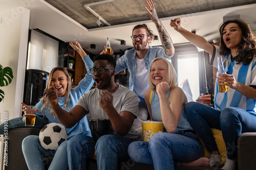 Excited group of people watching football, soccer sports match at home. Multi-ethnic group of emotional friend fans cheering for their favorite national team, drinking beer.  © lordn