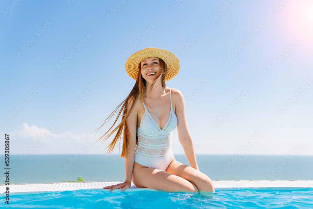 a slender woman in a white swimsuit in a straw hat sits on the pool.