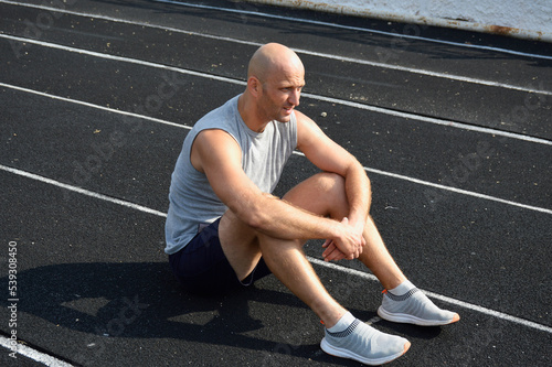 a young bald man in a gray jersey is resting after a workout on the rubber surface of the running track of the city stadium. active lifestyle, sports and physical education