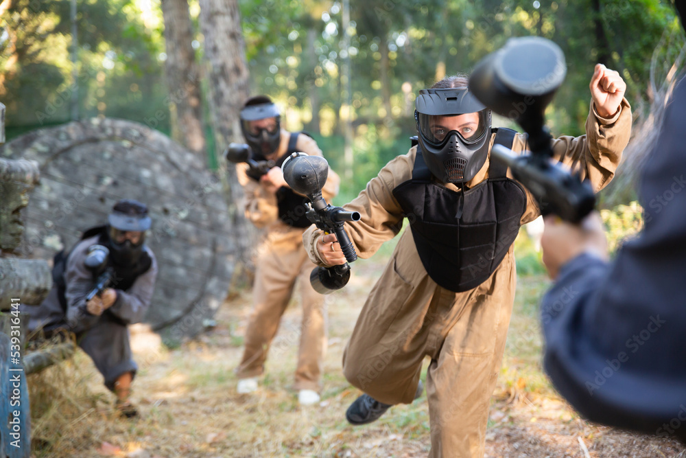 Fototapeta premium Young woman paintball player wearing protective clothes and helmet, rushing through enemy lines, shooting with paintball marker.