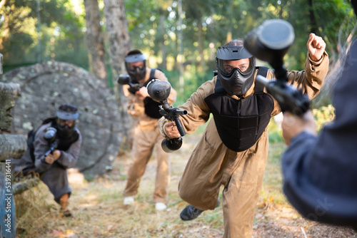 Young woman paintball player wearing protective clothes and helmet, rushing through enemy lines, shooting with paintball marker.