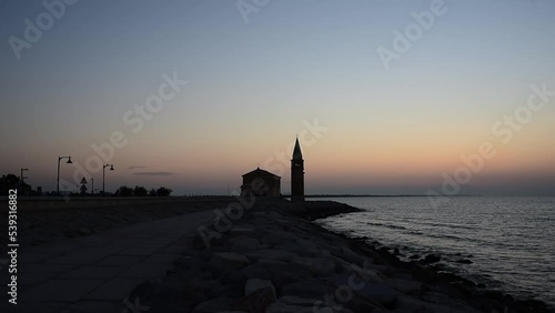 People stroll at dawn on the seafront of Caorle, in the background the sanctuary 