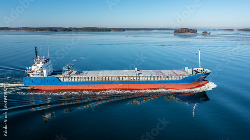 Aerial starboard side view of general cargo vessel making way ahead in Finnish archipelago during spring time. Glassy sea surface with few ice floes.