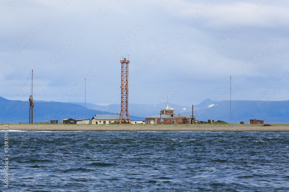 Lighthouse by the sea. Lighthouse tower and old buildings and structures on the shore. Marine transport infrastructure in the Far East of Russia. Spit Russkaya Koshka, Chukotka. Anadyr Bay, Bering Sea