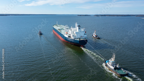 Tug boats escort large crude oil carrier through narrow Finnish archipelago. Ship's hull recently painted in dry dock. Aerial stern view.