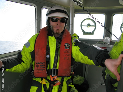 Work boat crew member in high visibility personal protective equipment inside small boat cabin. Person wearing SOLAS approved immersion suit and inflatable life jacket. photo