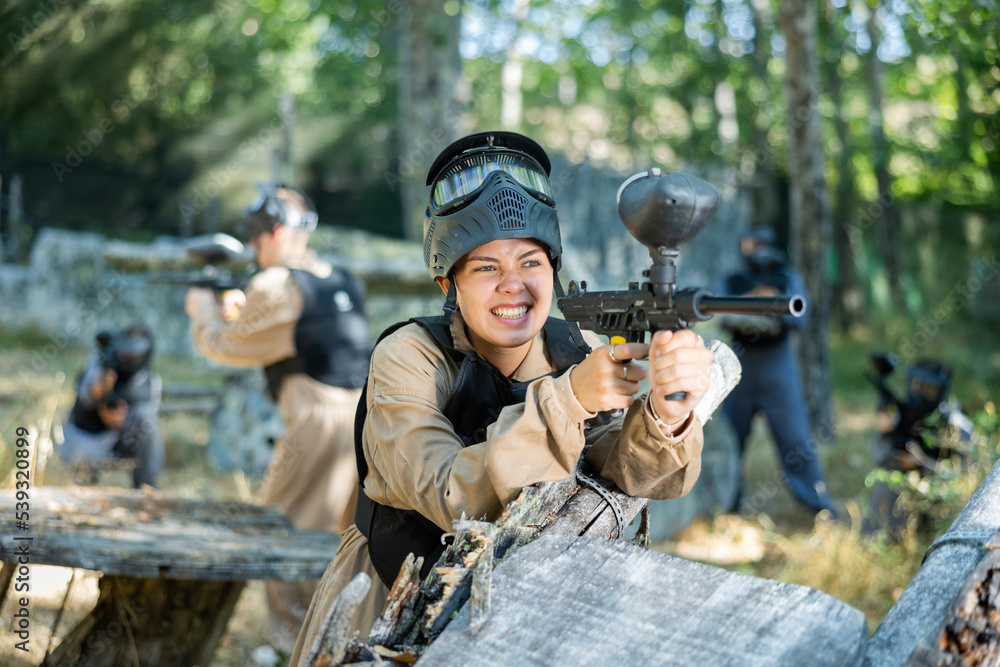 Fototapeta premium Happy europan girl standing behind barricade and targeting and shooting with paintball marker during training in forest venue.