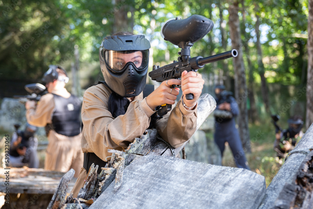 Fototapeta premium Girl in protective paintball outwear standing behind cover, aiming and shooting with paintball marker.