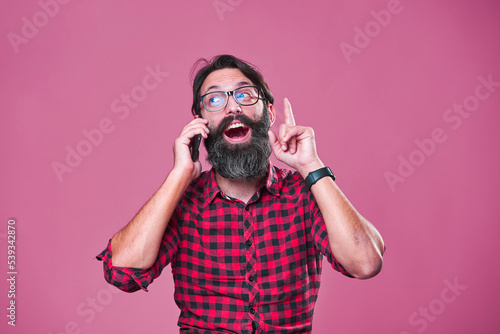 Bearded man with glasses coming up with solution, idea realization while talking on the phone; pink studio background photo