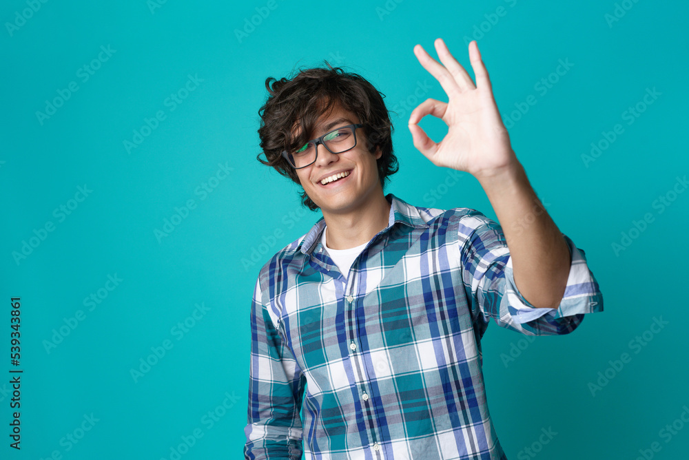 Young man with spectacles making an ok sign, expressing approval and looking cheerful and content; teal studio background Young man with spectacles making an ok sign, expressing approval and looking cheerful and content; teal studio background