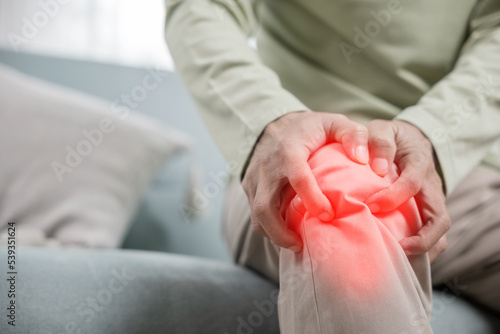Old man suffering from knee joint pain on sofa living room, bone pain in elderly at home, senior man knee problem painful, unhappy old age hand holding on knee pain after tendon surgery