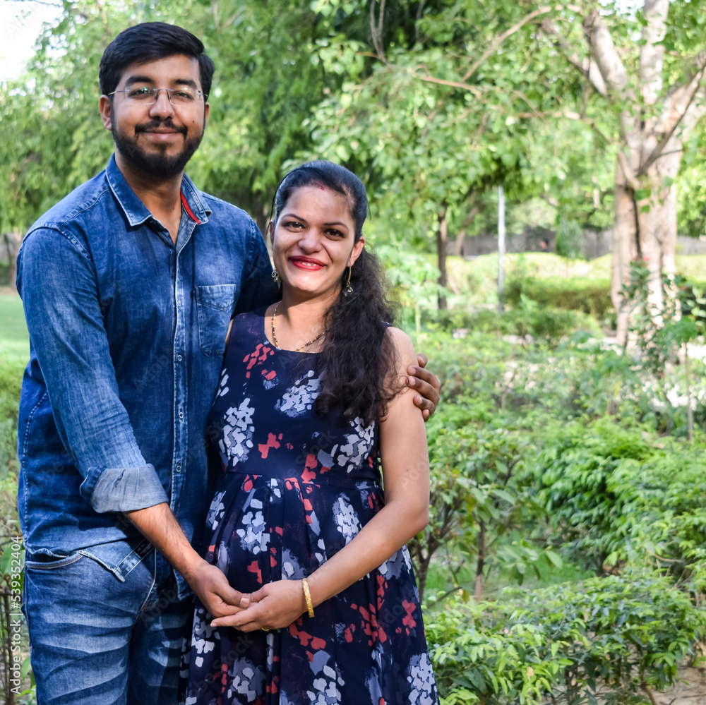 Indian couple posing for maternity baby shoot. The couple is posing in a lawn with green grass and the woman is falunting her baby bump in Lodhi Garden in New Delhi, India