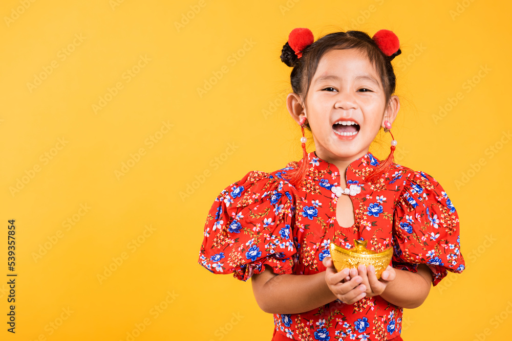 Chinese New Year. Happy Asian Chinese little girl smile wearing red cheongsam holding gold ingot, Portrait children in traditional dress hold golden bar, studio short isolated on yellow background Chinese New Year. Happy Asian Chinese little girl smile wearing red cheongsam holding gold ingot, Portrait children in traditional dress hold golden bar, studio short isolated on yellow background
