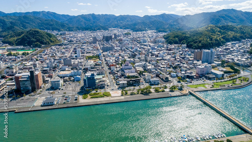 Photography 海岸沿いに広がる市街地と山々、海を隔てる堤防や橋を捉えた地方都市の空撮写真。青い海と街並み、背後に連なる緑豊かな山岳地帯の対比が特徴。地方自治体の観光PR、都市開発や不動産関連の資料、日本の風景を紹介する販促・広告用素材に。 | Aerial view of a coastal city with mountains in the background