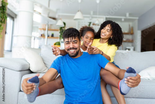 Happy African American dad and mom with excited proud daughter kid, playing flying superhero, reaching arm forward. Cheerful girl playing active game with family at home