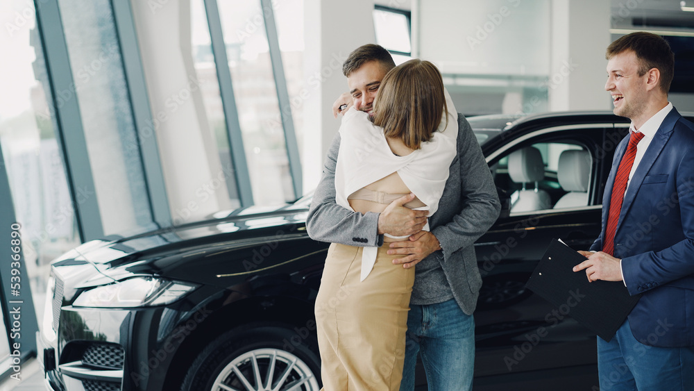 Young bearded man is buying car for his attractive wife, she is kissing and hugging him.