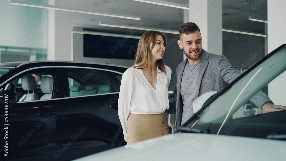 Attractive young couple confident bearded guy and his smiling pretty girlfriend are choosing new automobile together looking at luxury car in auto dealership and talking.