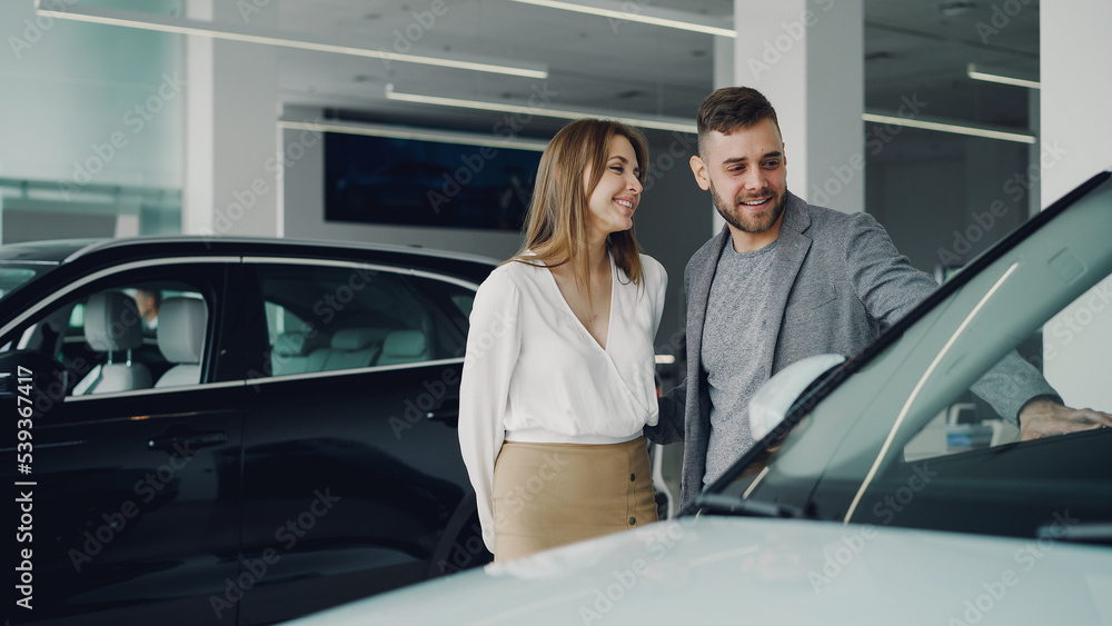 Attractive young couple confident bearded guy and his smiling pretty girlfriend are choosing new automobile together looking at luxury car in auto dealership and talking. Attractive young couple confident bearded guy and his smiling pretty girlfriend are choosing new automobile together looking at luxury car in auto dealership and talking.