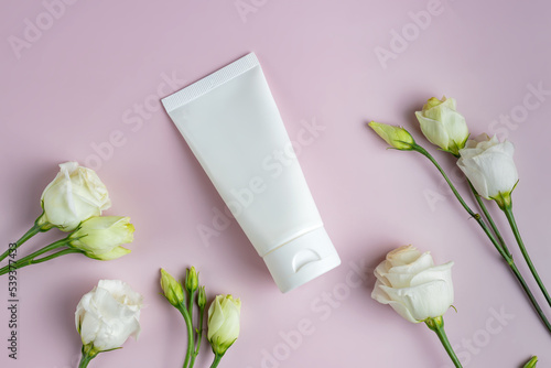Blank plastic white tube for cream, mask, shampoo or toothpaste on pink background among eustoma flowers. Spring presentation of cosmetics package. Mockup