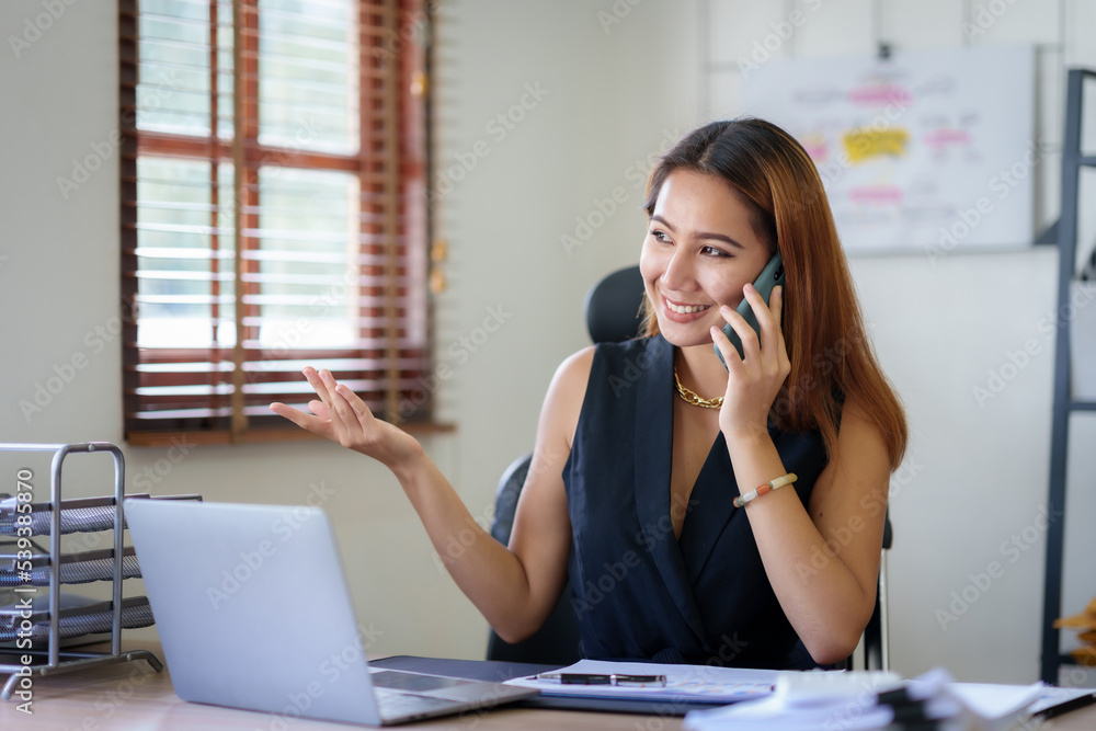 Happy Asian businesswoman smiling and talking on the phone with a client in the office with a bunch of papers on the desk. Happy Asian businesswoman smiling and talking on the phone with a client in the office with a bunch of papers on the desk.