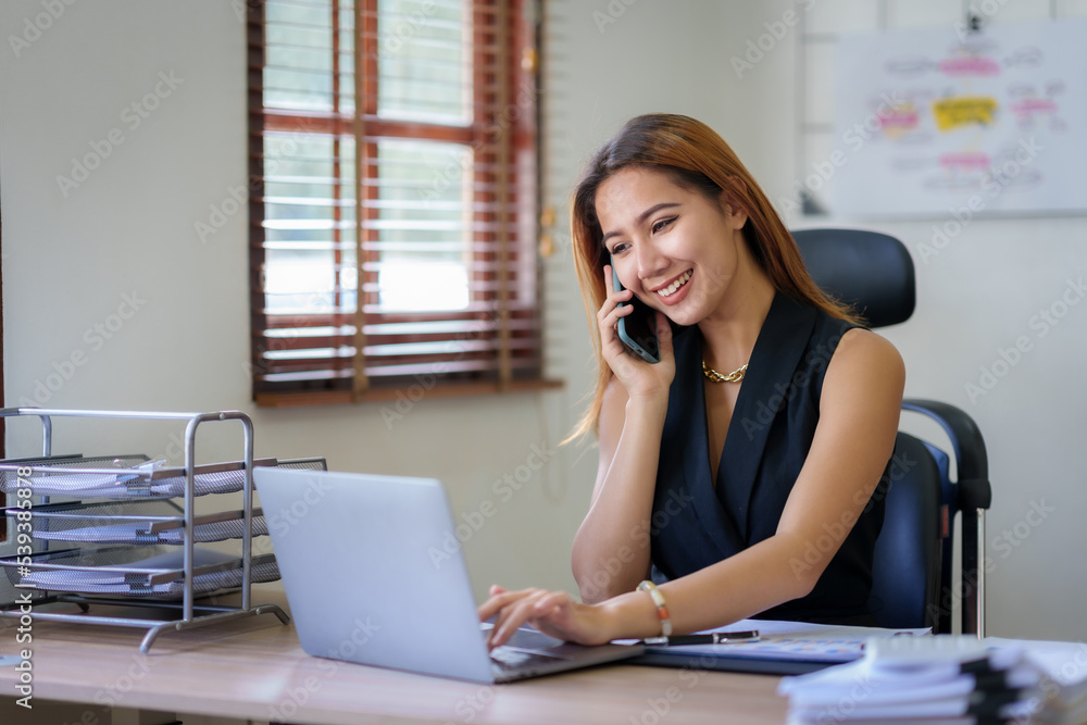 Happy Asian businesswoman smiling and talking on the phone with a client in the office with a bunch of papers on the desk. Happy Asian businesswoman smiling and talking on the phone with a client in the office with a bunch of papers on the desk.