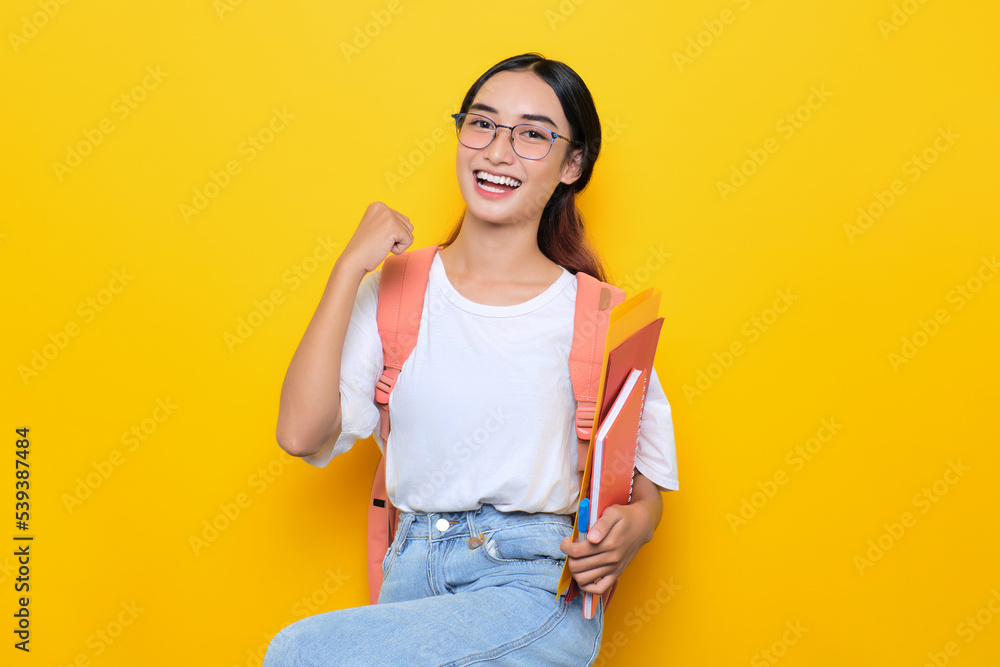 Excited pretty young student girl wearing backpack and eyeglasses holding books, raises fist in excitement isolated on yellow background Excited pretty young student girl wearing backpack and eyeglasses holding books, raises fist in excitement isolated on yellow background