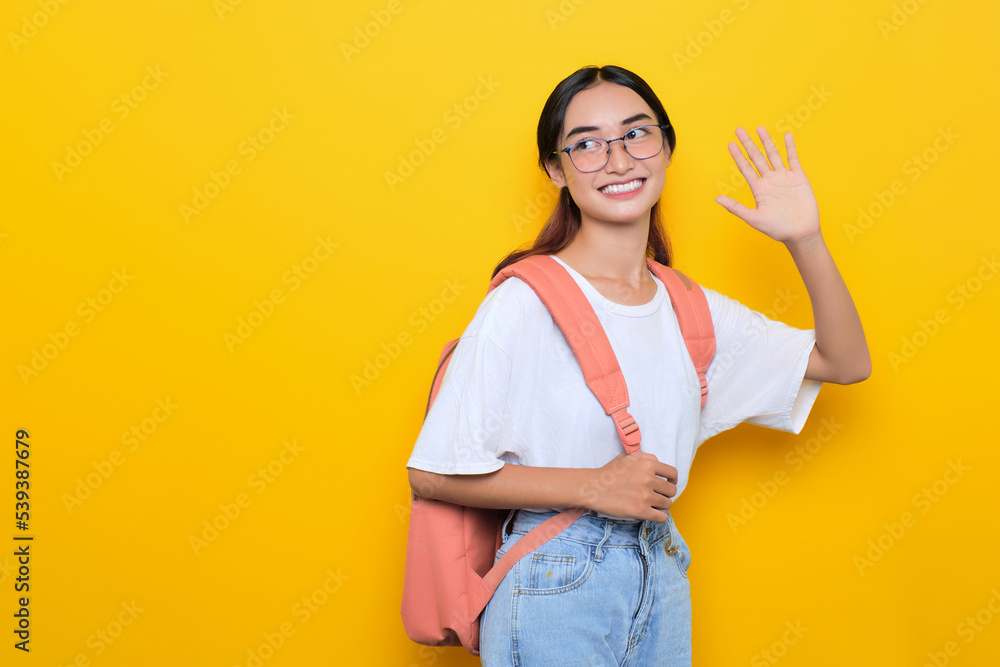 Cheerful pretty young student girl wearing backpack and eyeglasses waving hand say hello isolated on yellow background Cheerful pretty young student girl wearing backpack and eyeglasses waving hand say hello isolated on yellow background