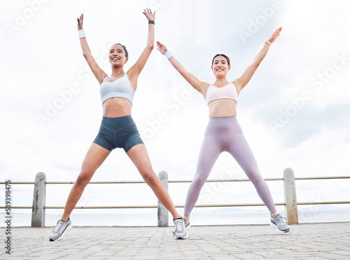 Jumping jack, fitness and women training cardio, happy with workout and smile during exercise in the city by the ocean. Young friends with energy for sports collaboration and stretching in the street photo