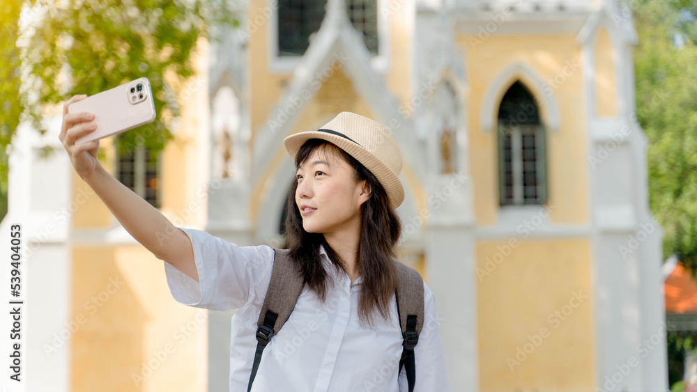 Young Asian woman traveler wearing white shirt and hat making picture on smartphone, happy female tourist posing for selfie using mobile phone camera