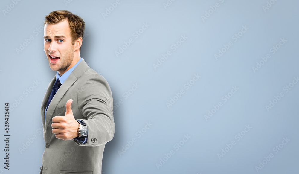 Excited businessman showing thumbs up like hand sign gesture, in grey confident suit, blue tie, isolated over grey background. Handsome happy man. Copy space area for ad slogan, advertising text. Excited businessman showing thumbs up like hand sign gesture, in grey confident suit, blue tie, isolated over grey background. Handsome happy man. Copy space area for ad slogan, advertising text.