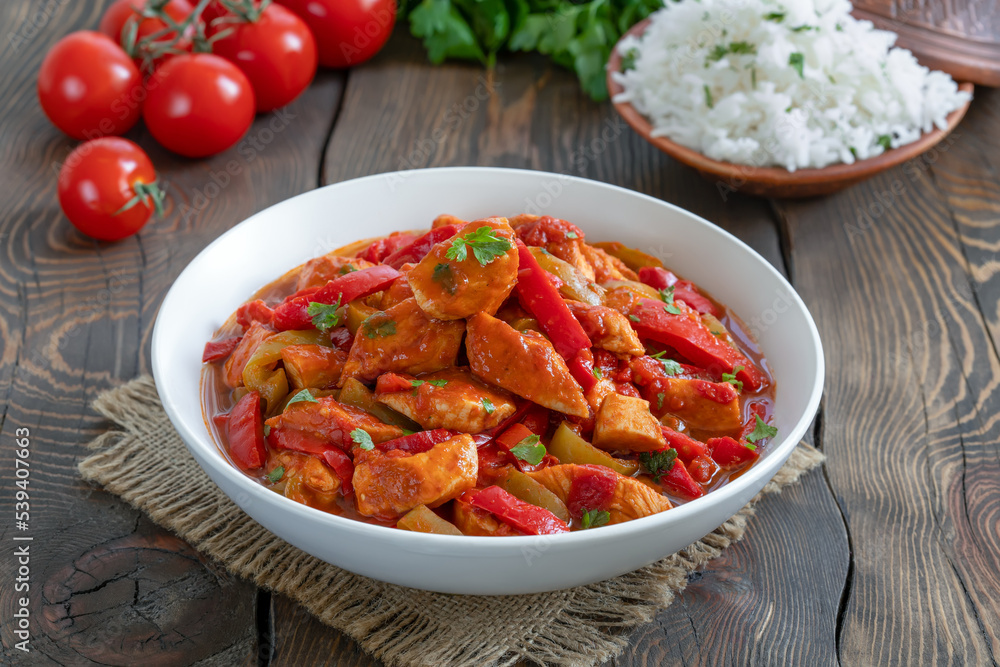 Tomato chicken stew. Braised chicken breast in tomato sauce mixed with onion, red and green bell pepper served with bowl of cooked rice on background. Tomato chicken stew. Braised chicken breast in tomato sauce mixed with onion, red and green bell pepper served with bowl of cooked rice on background.