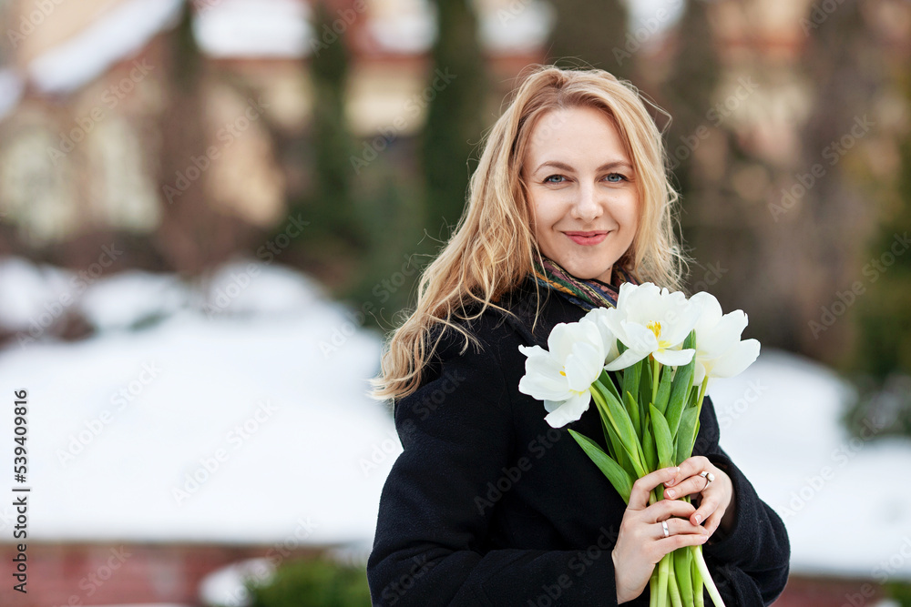 Fototapeta premium Elegant smiling young woman with long blonde hair in a black coat poses in the city. Portrait of a beautiful modern woman with flowers
