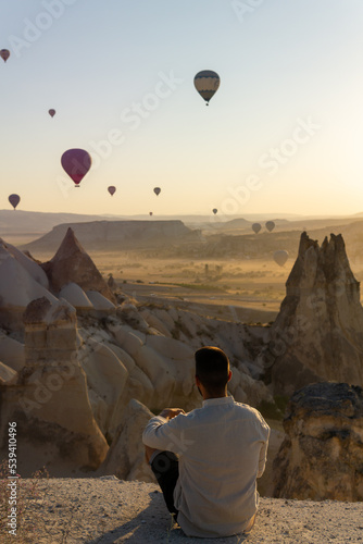 Unrecognizable tourist sitting in the foreground enjoying views of the valley of love and hot air balloons flying at sunrise in Cappadocia, Turkey.