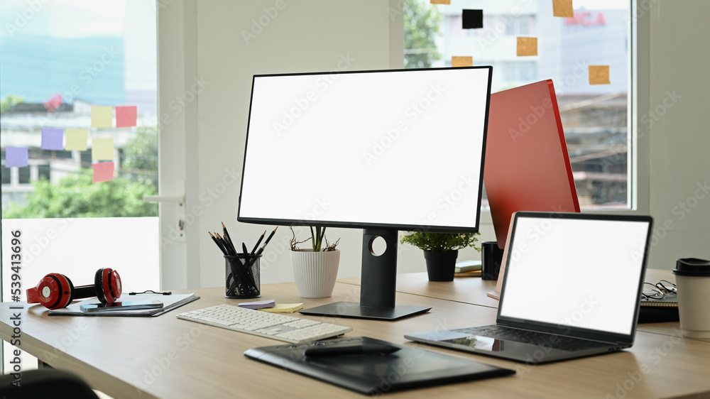 Creative office studio interior with laptop, blank screen computer, graphic tablet and stationery on wooden table Creative office studio interior with laptop, blank screen computer, graphic tablet and stationery on wooden table