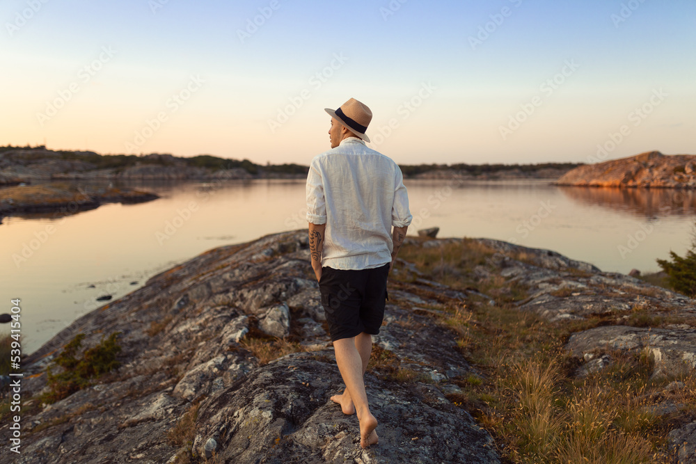 A caucasian man with straw hat and a white linnen shirt walking on a rock by the sea at sunset.