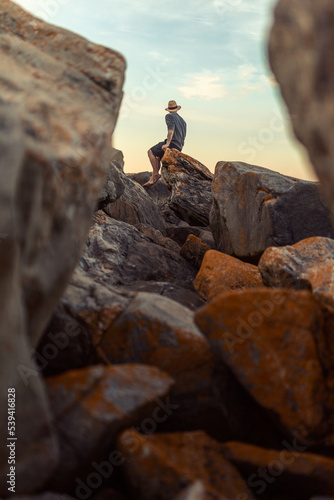 A man wearing a straw hat and shorts sitting on rocks at sunrise. © Francis