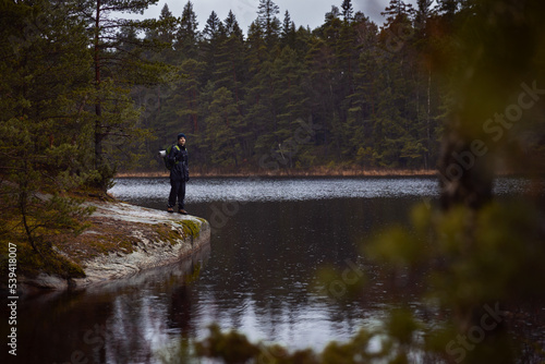 A caucasian man with a backpack standing on a rock by a lake in a forest on a rainy day.