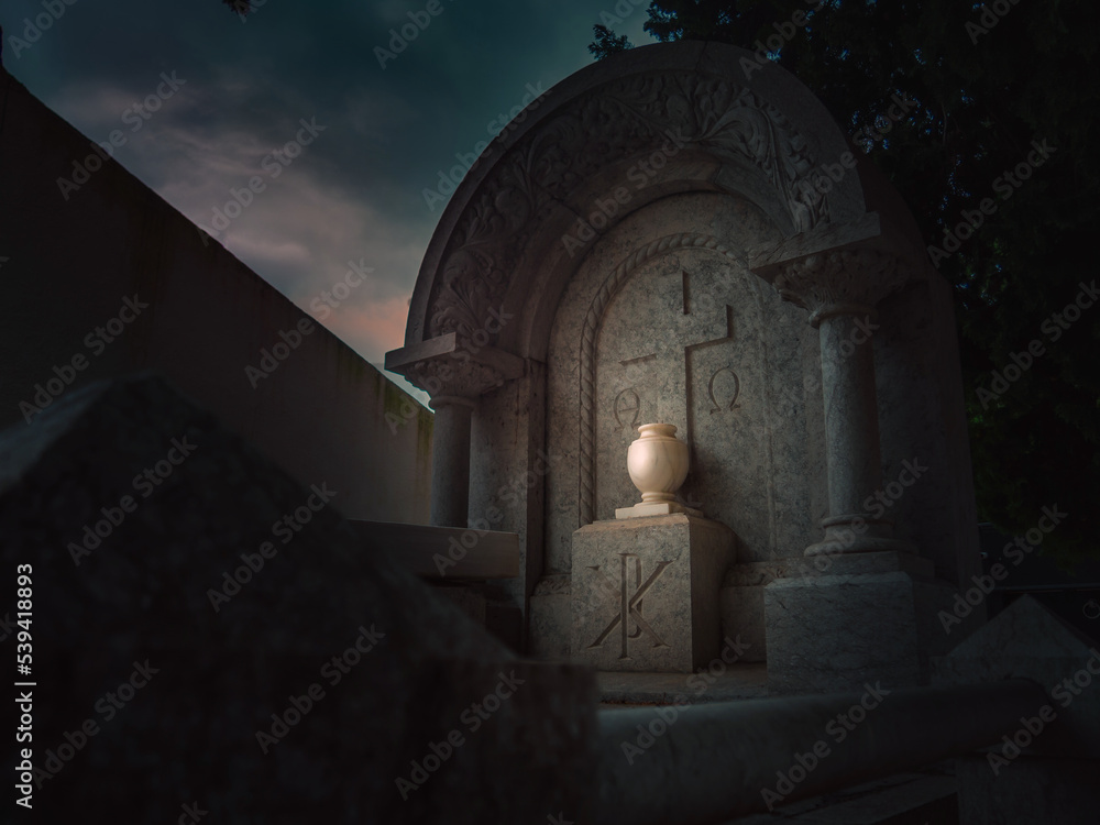 Fototapeta premium Low light dramatic and smooth red tinted tomb in a obscure Christian cemetery adorned with a carved floral design with an urn illuminated by a beam of light at night in Vandellòs, Catalonia, Spain
